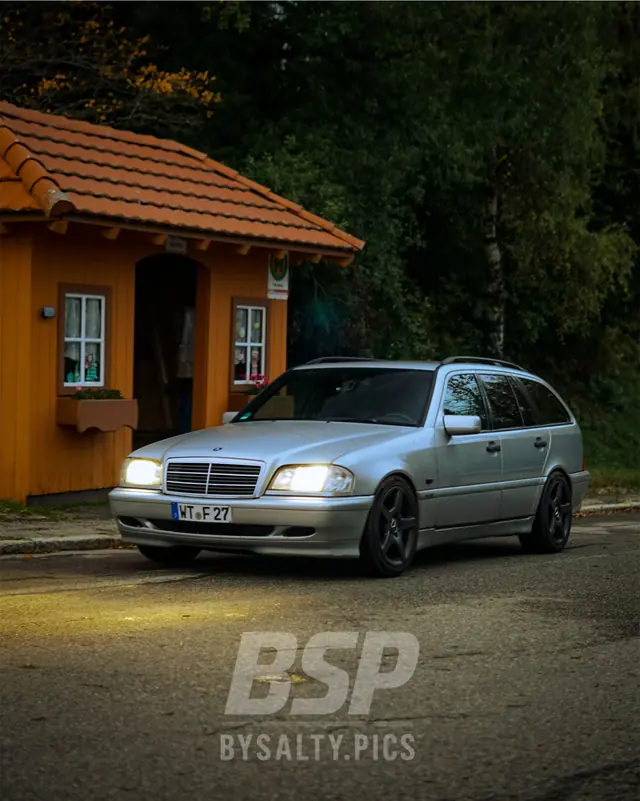 Silberner Mercedes-Benz C-Klasse T-Modell vor einem orangefarbenen Holzhäuschen im Wald, Automobilfotografie